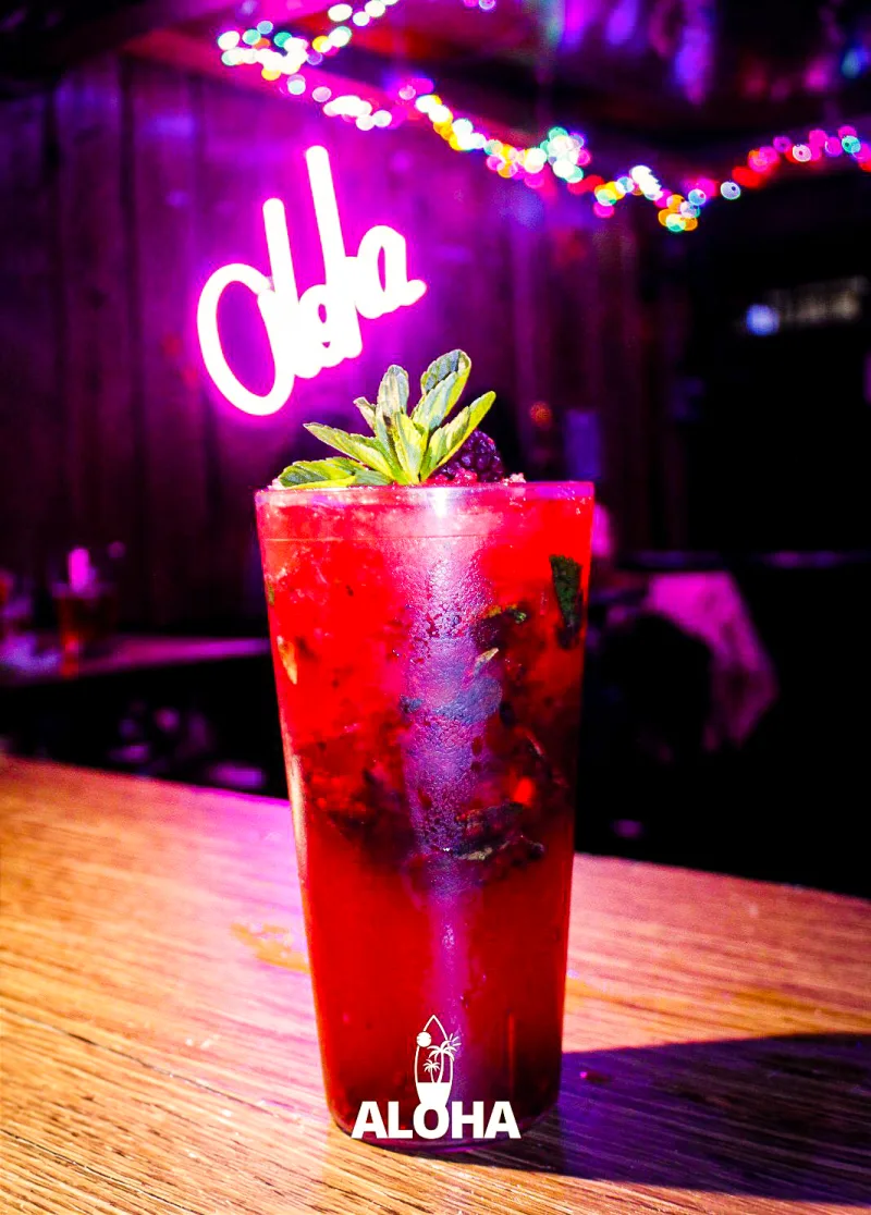 Coctel rojo carmesí con menta y neón rosa Aloha al fondo